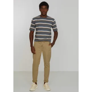 Man standing against a light background wearing a blue, beige, and white striped short-sleeve T-shirt with tan chinos and cream sneakers; hands in pockets.