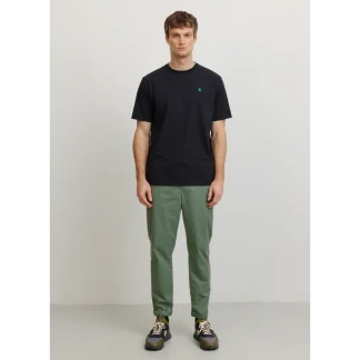 Man standing against a light wall, wearing a black short-sleeve T-shirt with a small green logo and olive-green pants, facing forward.